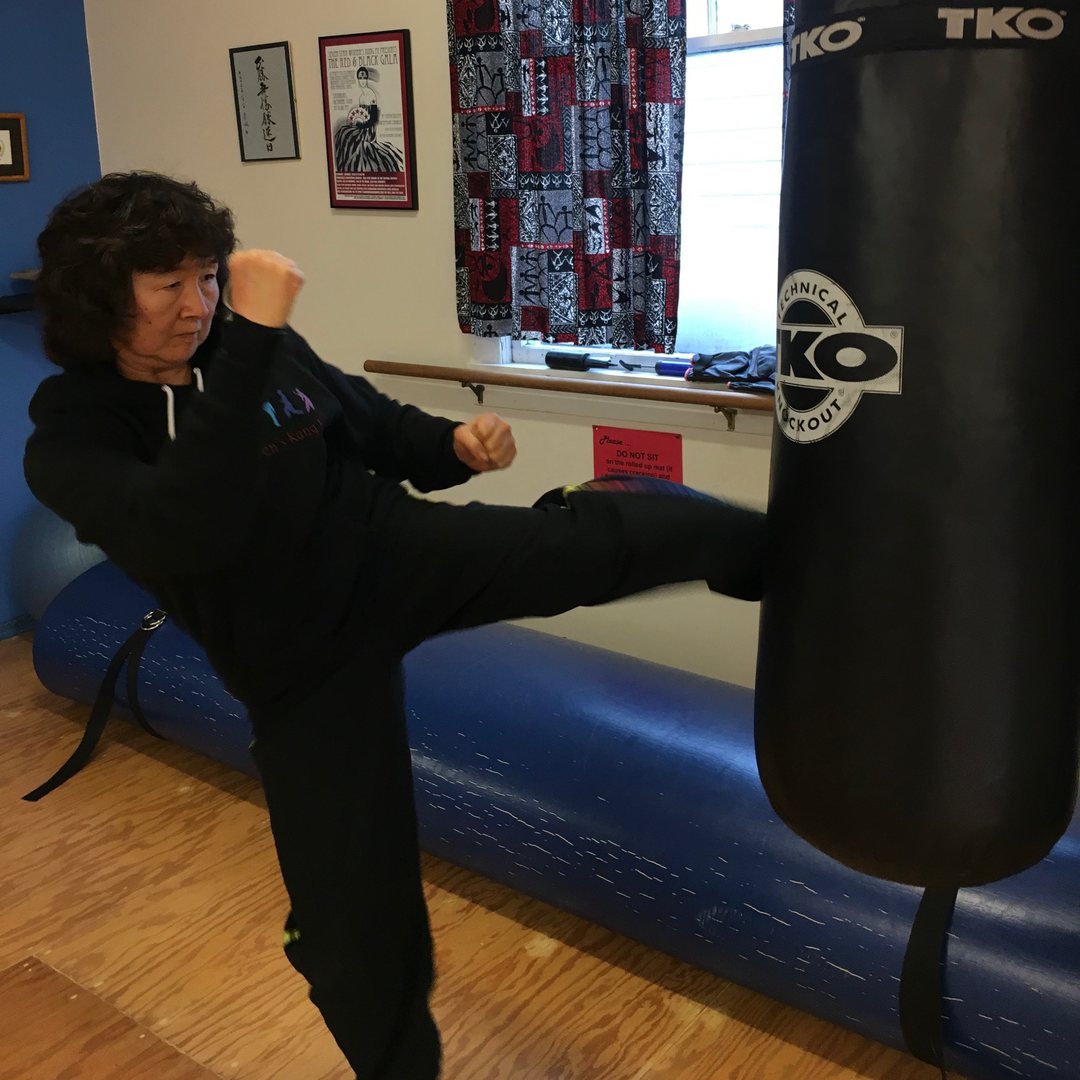 Mavis Tsai practicing Kajukenbo, mid-kick against a heavy bag in a martial arts studio.