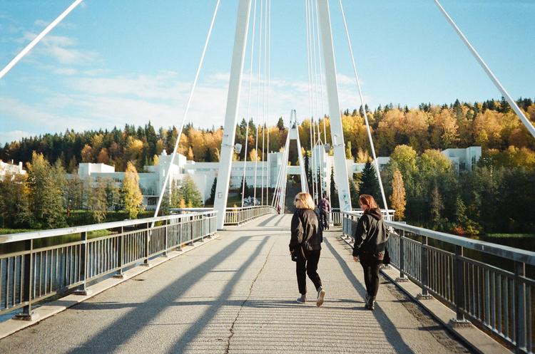 Two people walking across a pedestrian bridge toward autumn trees and white buildings.